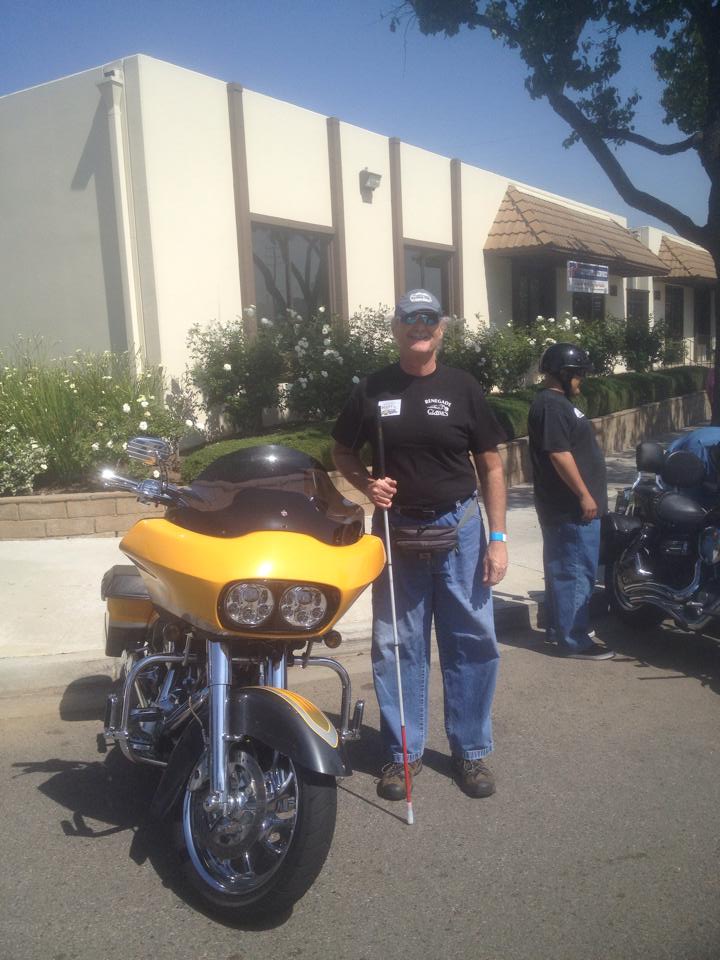 Man with a cane standing beside a yellow motorcycle outdoors.