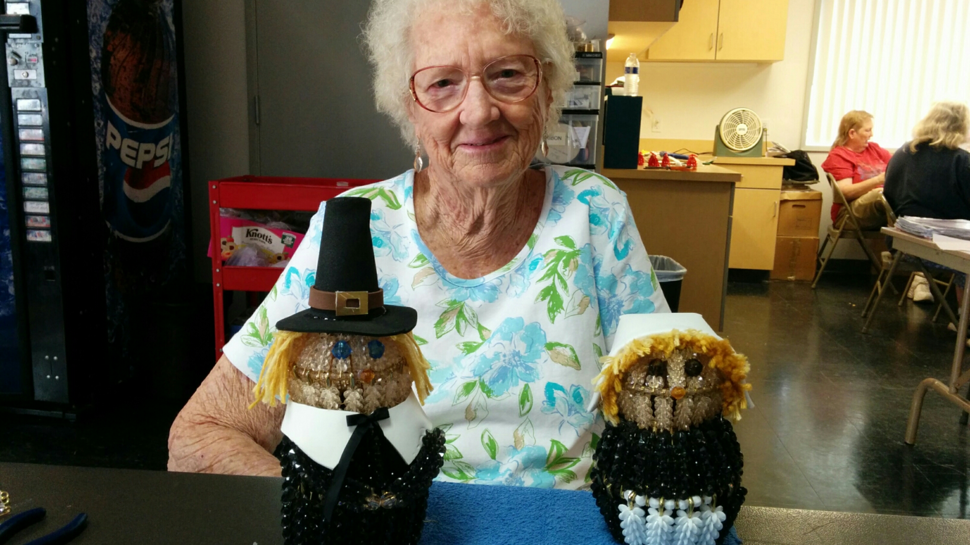 Elderly woman smiling with two festive Pilgrim-themed scarecrows.