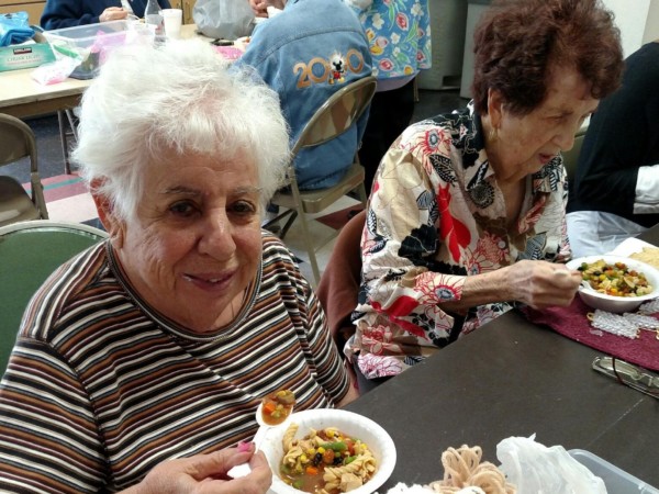Elderly woman smiling and holding a bowl of food at a gathering.