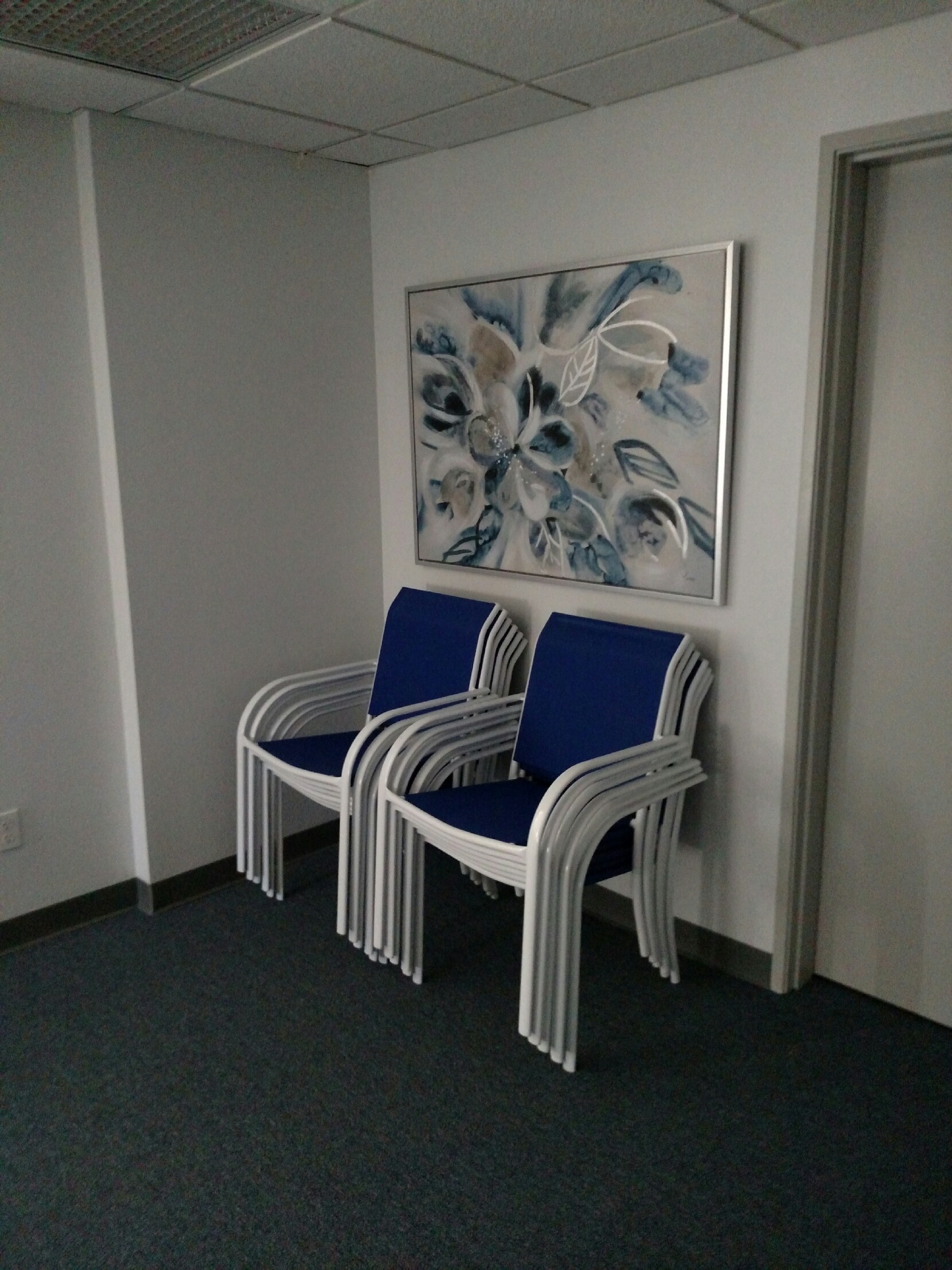Stacked blue and white chairs under a large floral painting in a simple room.