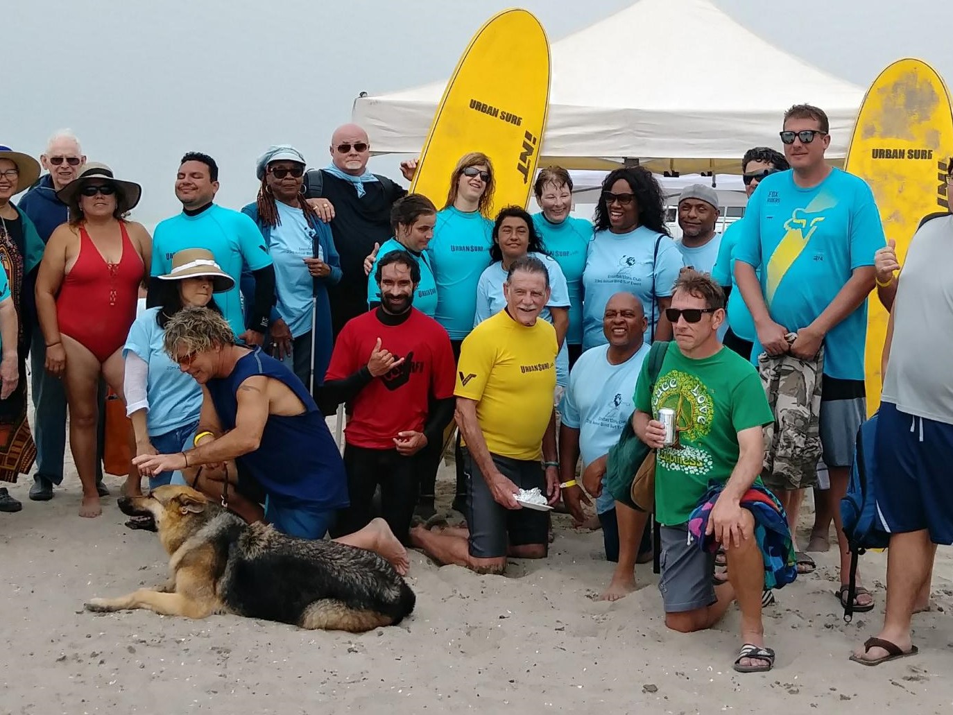 Group of people and a dog posing on a beach with a yellow surfboard.