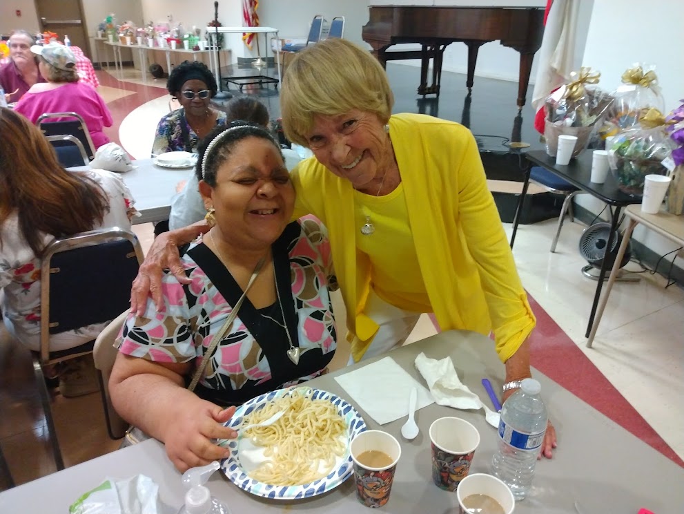 Two women smiling warmly at a gathering with food and drinks.